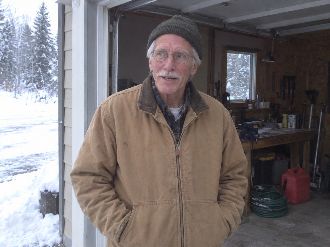Gene Halvorsen in a knit cap and tan work jacket, standing in his garage doorway with snow on the ground outside.